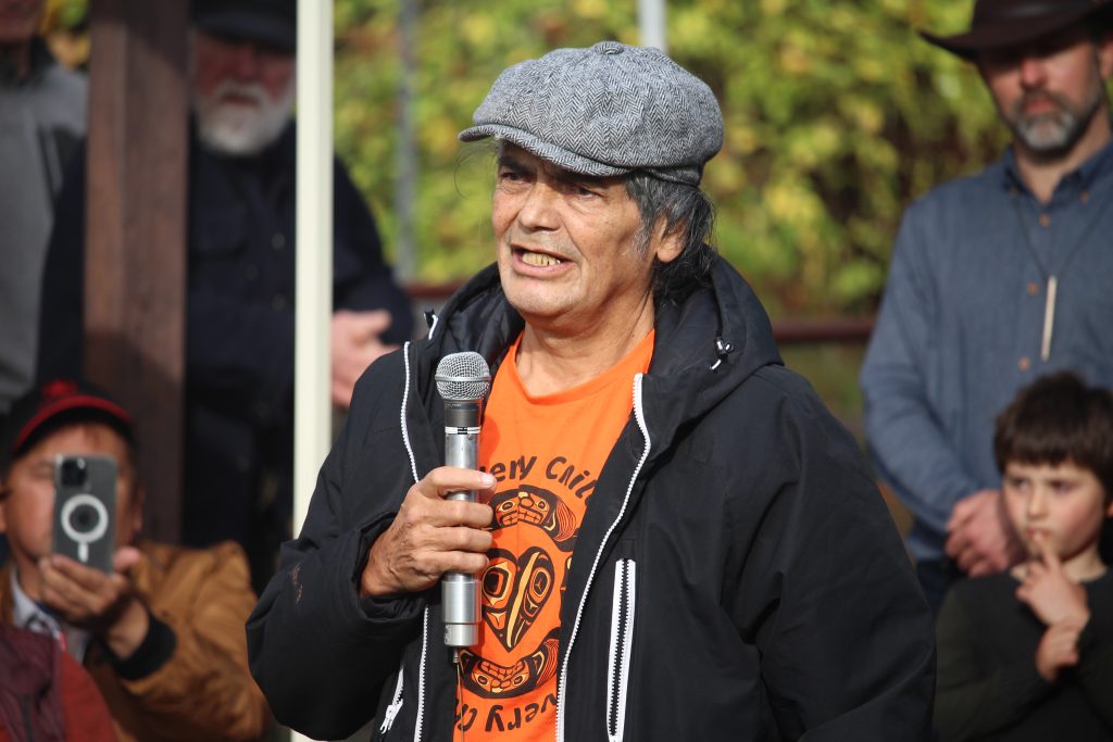 Elder Charles Joseph holds a microphone and speaks. He is wearing an orange "every child matters" shirt and is wearing a grey cap and black zip-up jacket.