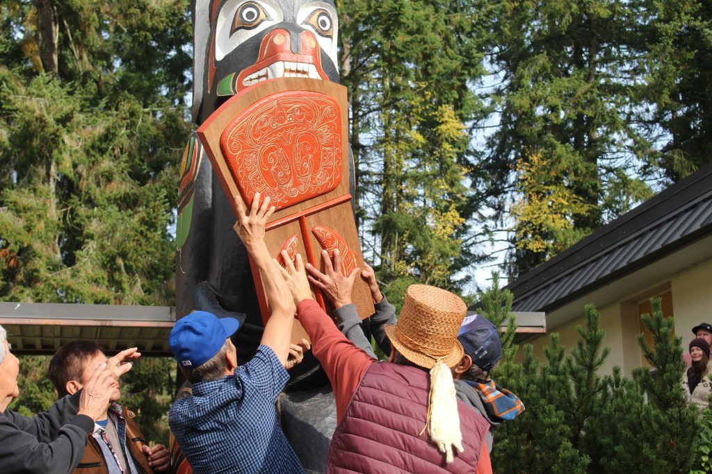 Photo taken from behind as Rupert Scow (centre and wearing a Cedar hat) works with his brothers on either side to raise a copper shield and insert it in the hands of the bear on the totem pole.