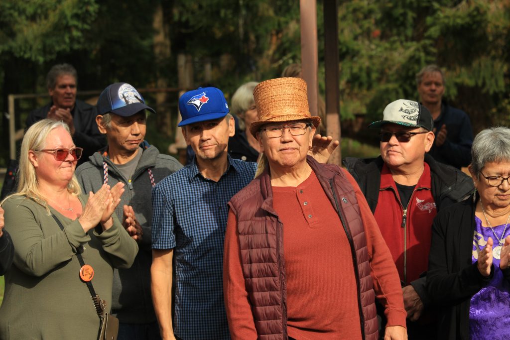 Rupert Scow stands in the middle of the photo, wearing a cedar hat. Behind him and on both sides of him are friends and family clapping. His brother, behind him, reaches out to put a hand on Rupert's shoulder.